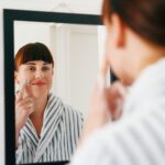 Femme appliquant une crème hydratante devant un miroir dans une salle de bains.